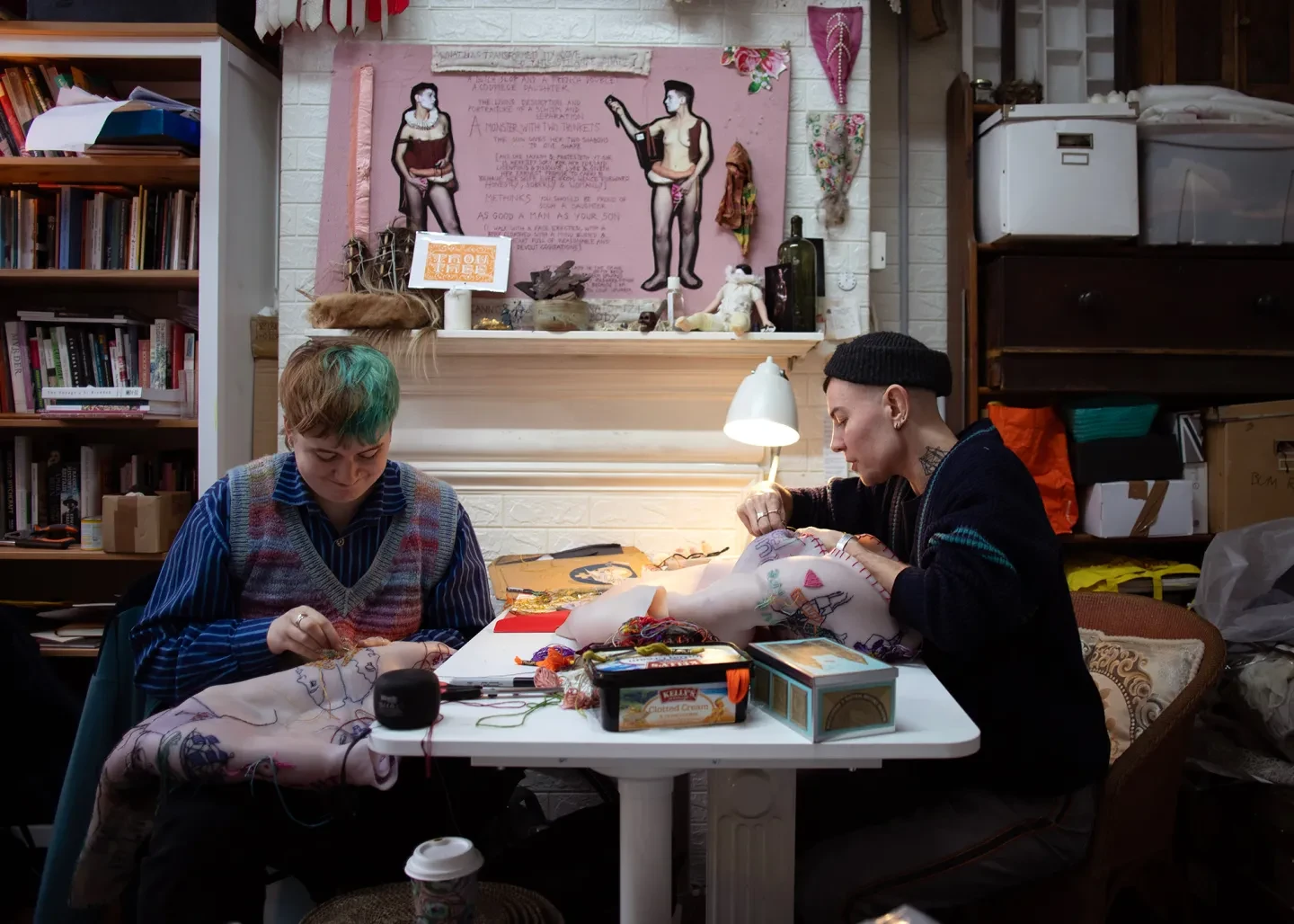 Two people sit sewing at a table, surrounded by bookshelves, boxes of crafting equipment and a display wall with reference images for their work